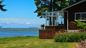 Uma casa com uma estufa de vidro com vista para um lago, um deck de madeira e um jardim florido.