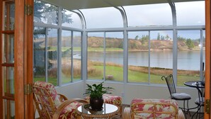 Uma sala de sol com grandes janelas com vista para um lago, móveis de vime e uma planta em vaso.