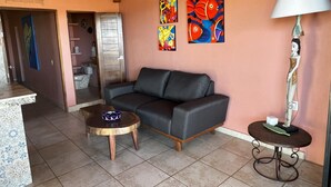 A living room with a sofa, a coffee table, and a lamp. There are paintings on the wall and a door leading to another room.