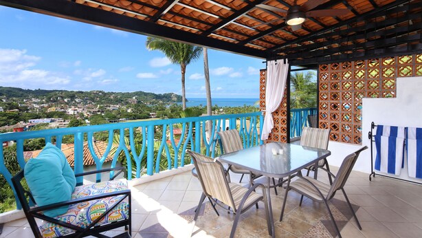 A covered outdoor seating area with a table and chairs, a view of the sea, and a decorative fence.