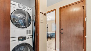 A laundry room with stacked washers and dryers, a wooden door, and a hallway leading to a bedroom with a bed.