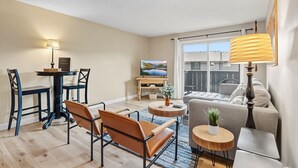 A modern living room with a sofa, coffee table, and chairs. There is a dining table with chairs, a TV, and a large window with a view of buildings.