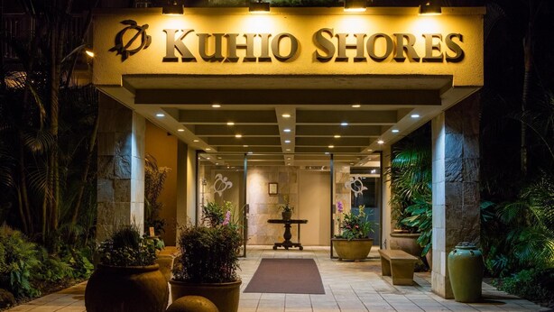 La entrada a KuHio Shores, con fachada de piedra, plantas en macetas y un camino bien iluminado que conduce al interior.