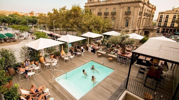 A rooftop pool area with outdoor seating and umbrellas, surrounded by trees and a historic building in the background. - Oasis Hotel (Barcelona)