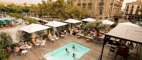 A rooftop pool area with outdoor seating and umbrellas, surrounded by trees and a historic building in the background.