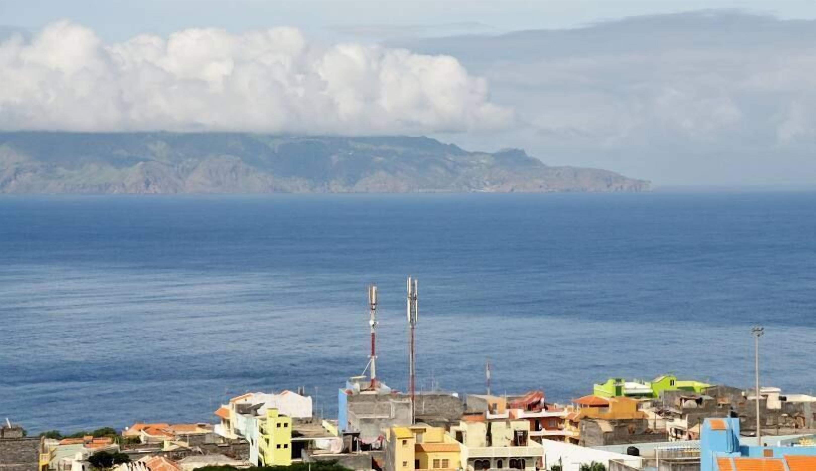 Un poble costaner amb edificis a prop del mar, un far i un paisatge de muntanyes al fons.