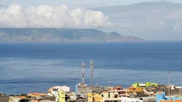 Un poble costaner amb edificis a prop del mar, un far i un paisatge de muntanyes al fons.
