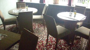 A dining area with two round tables, chairs, a patterned carpet, and a window with curtains. - Hotel Europa (Holsthum)