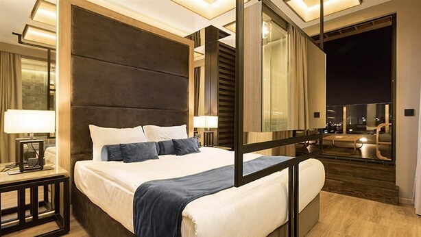 A modern hotel room with a large bed, a nightstand, and a view of the city at night. - Eden Garden Suites (Belgrade)