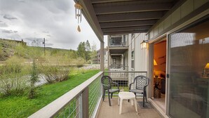 A balcony with a view of a river and greenery, two chairs, and a table. - Hi Country Haus Unit 1903 River view from private deck 2 bedroom modern interior (Winter Park)