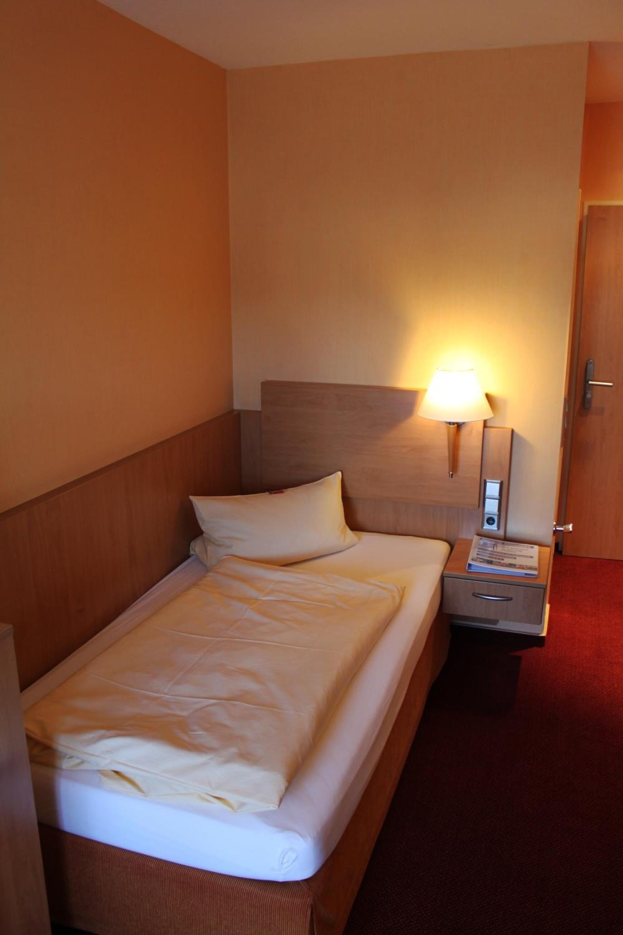 Ein Hotelzimmer mit einem Einzelbett, einem Nachttisch mit Lampe und einer Tür.