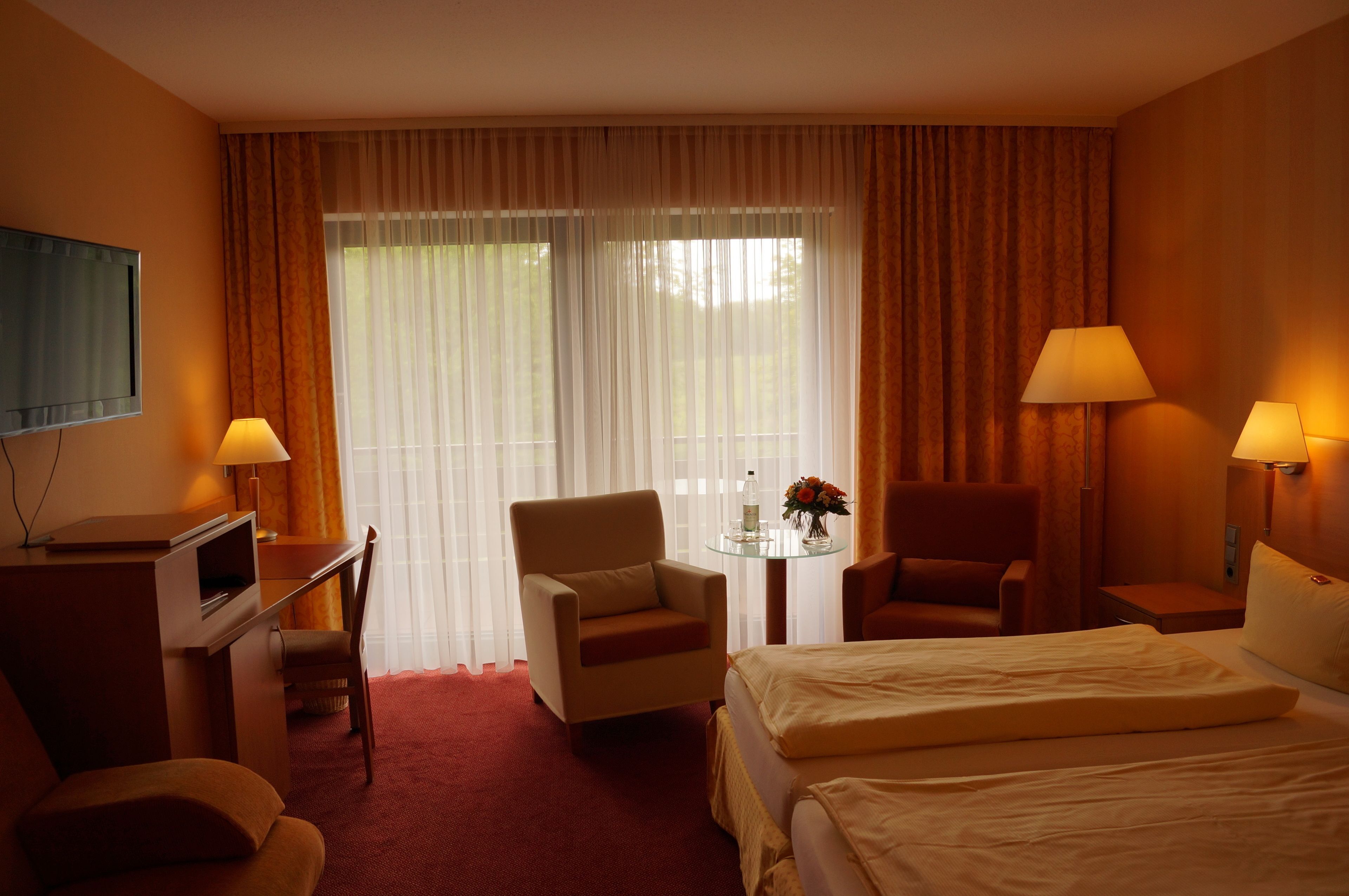 A hotel room with a bed, two armchairs, a desk with a television, a small table with a vase, and curtains.
