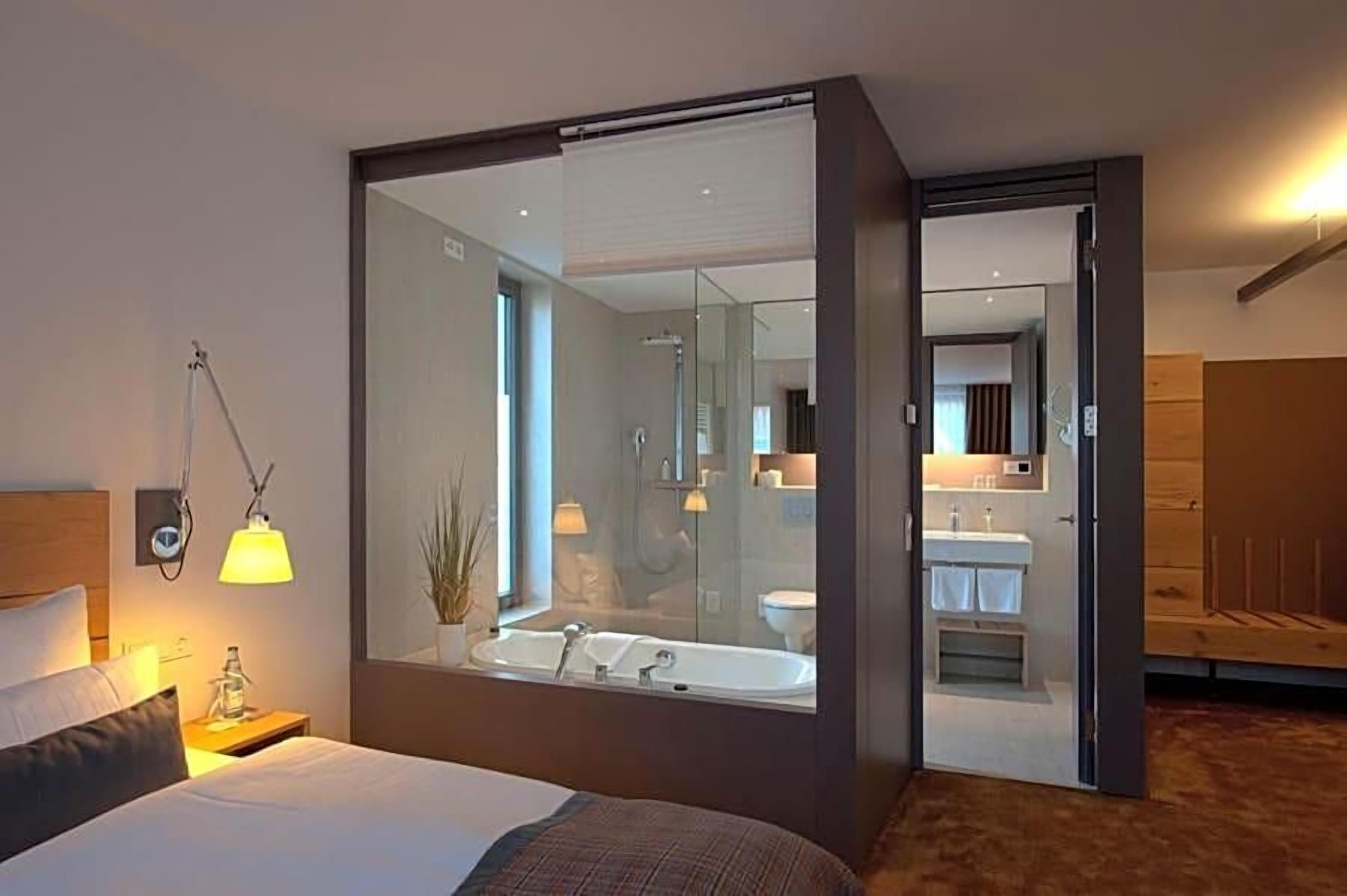 Ein modernes Hotelzimmer mit einem großen Bett, einem Nachttisch mit Lampe, einem Spiegel und Blick ins Badezimmer.
