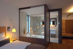 A modern hotel room with a large bed, a bedside table with a lamp, a mirror, and a view into the bathroom.