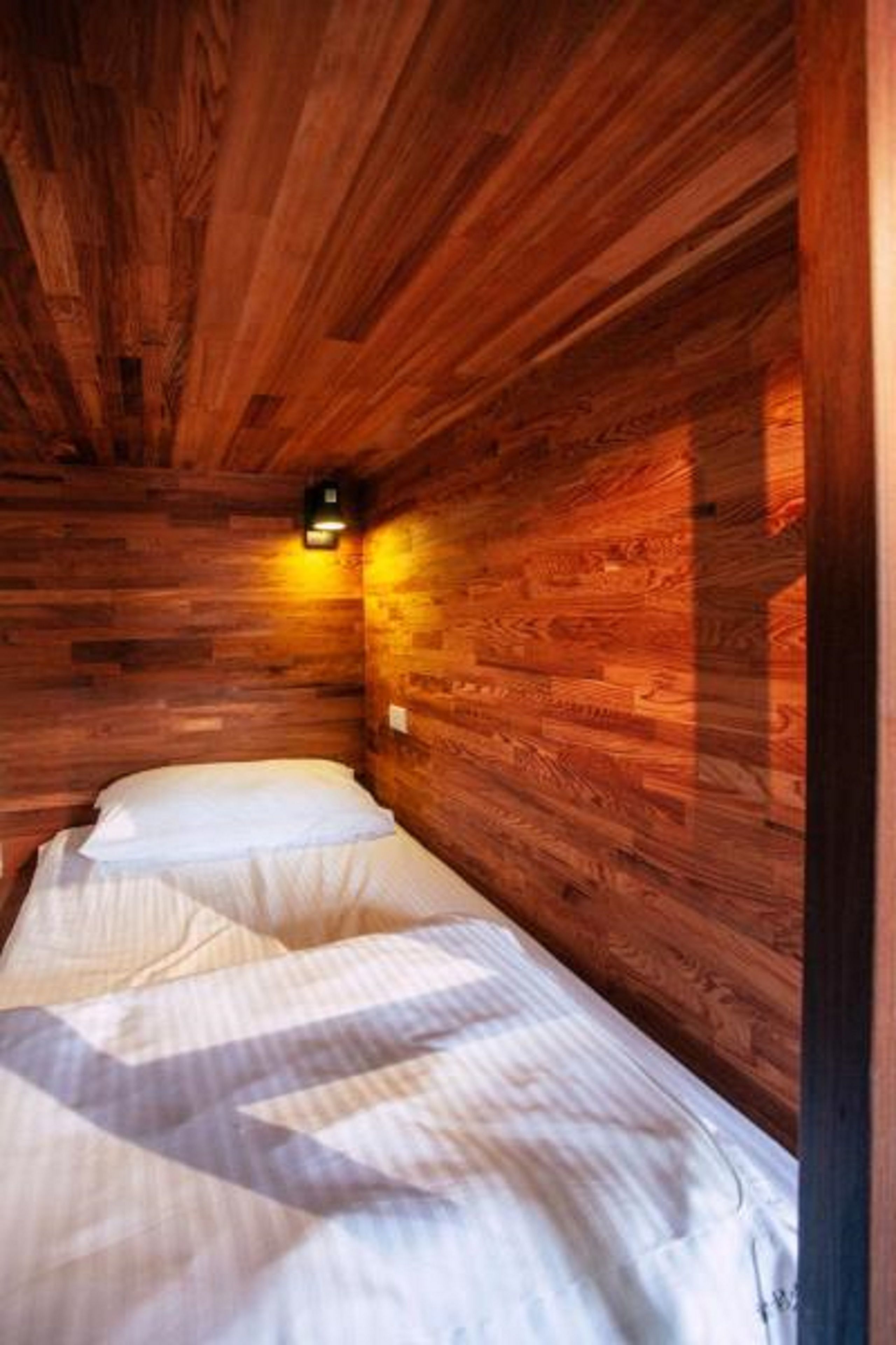 A wooden cabin room with a bed, a wall-mounted light, and a window.