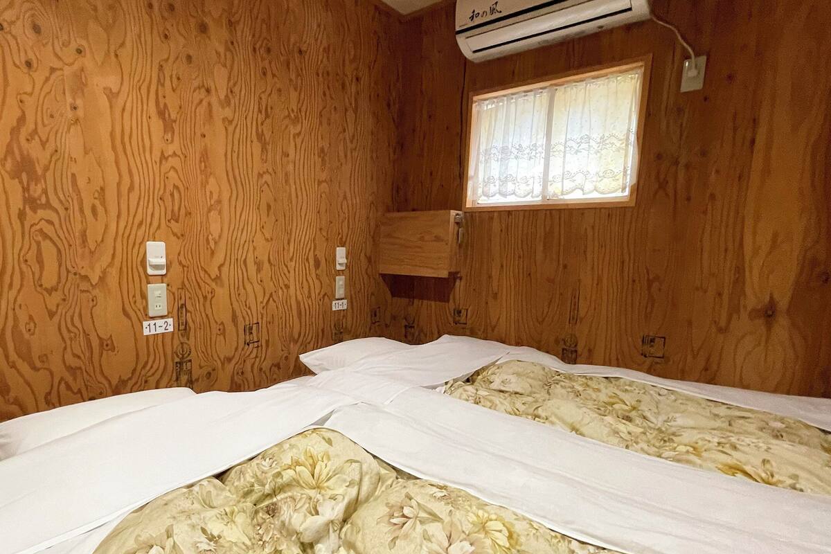 A wooden room with two beds, a window with lace curtains, and an air conditioning unit.
