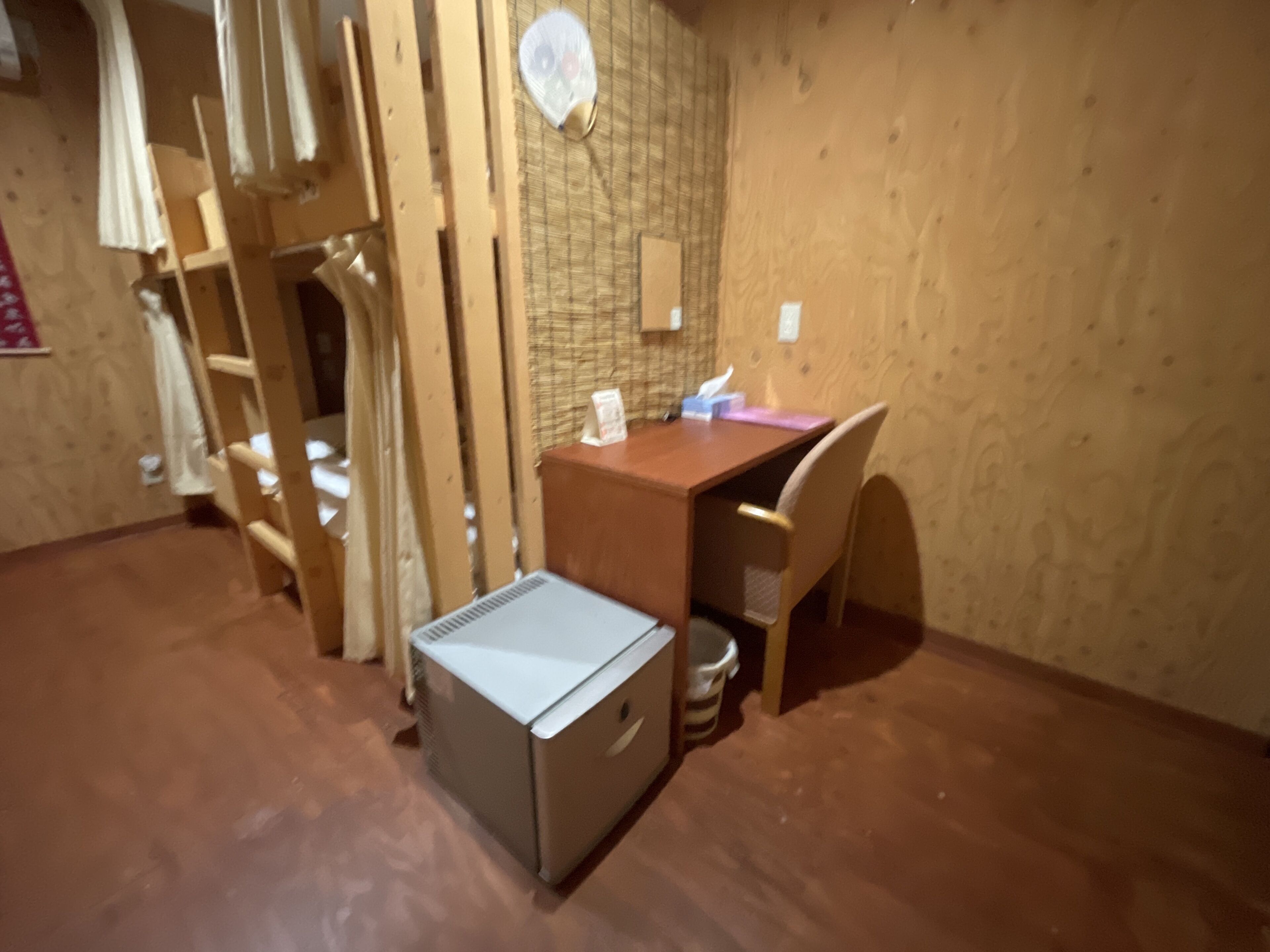 Quarto compacto com uma pequena escrivaninha, cadeira e um refrigerador compacto.