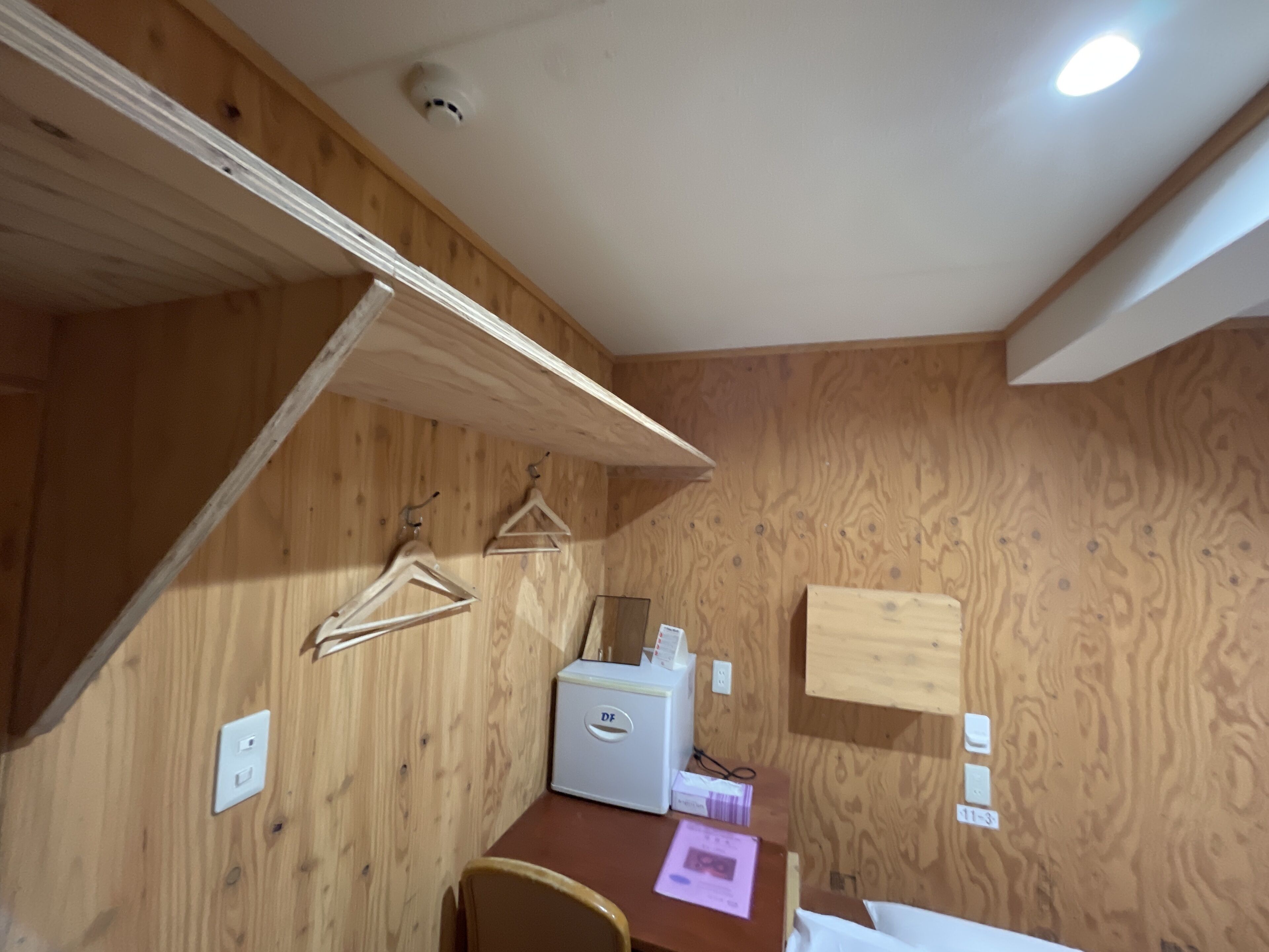 Ein Zimmer mit Holzwänden, einem Schreibtisch, einem Stuhl und einem kleinen, weißen Kühlschrank.