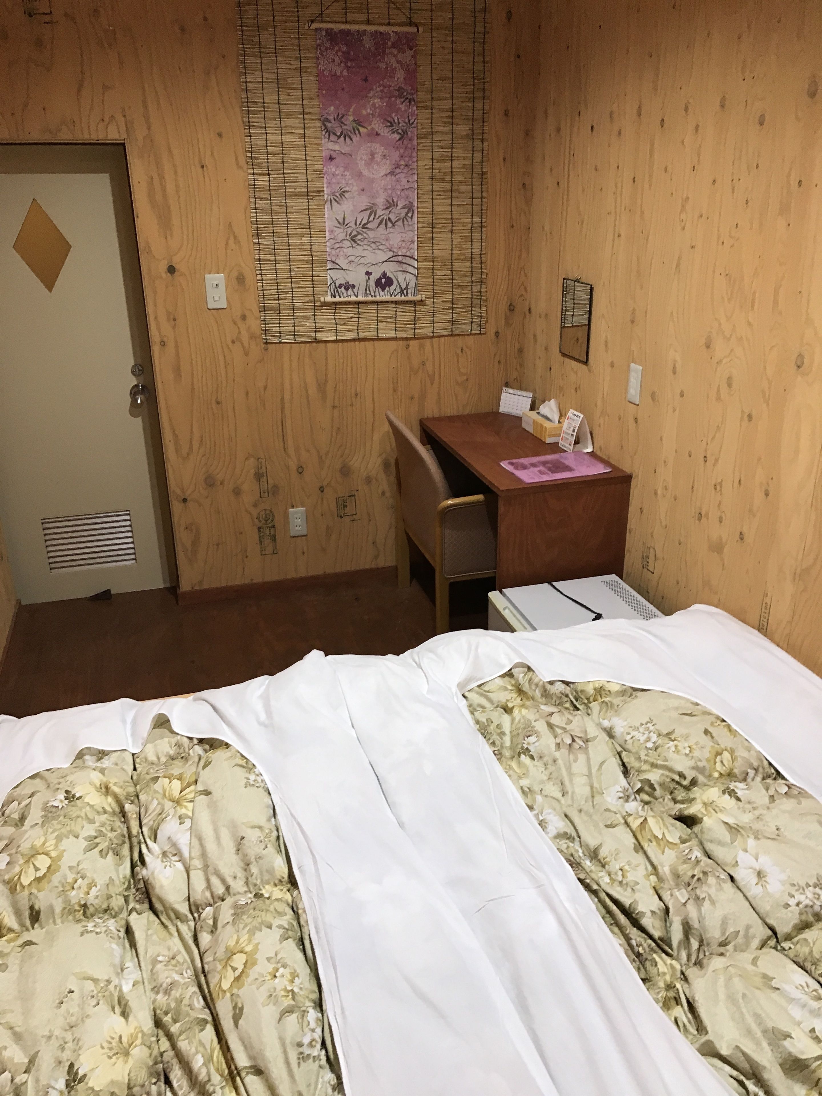 A hotel room with a wooden wall, a bed with floral bedding, a desk with a chair, and a calendar on the wall.