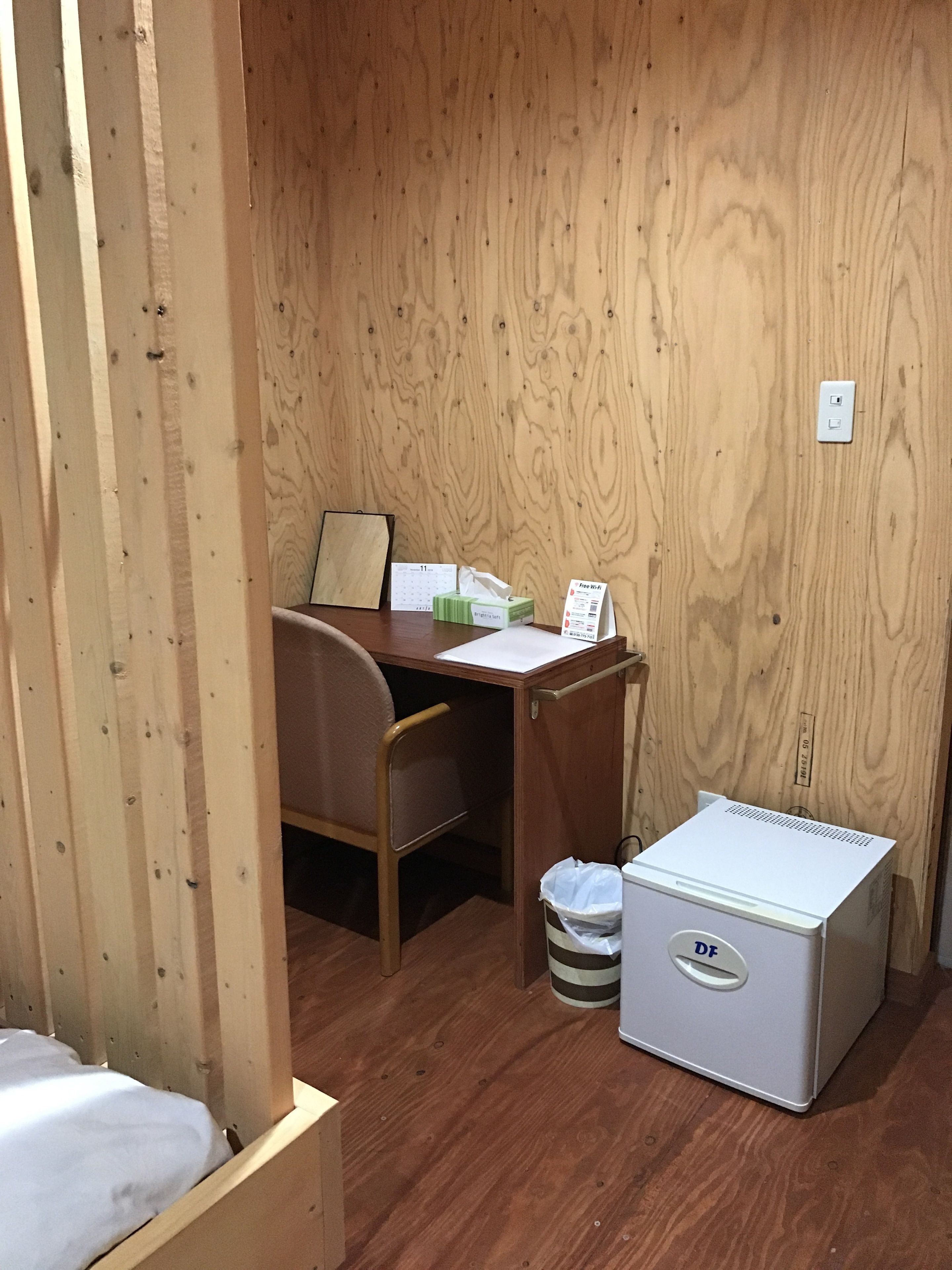 A small, compact room with a wooden wall, a desk with a chair, a bed, and a small white refrigerator.