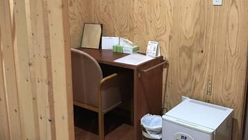 A small, compact room with a wooden wall, a desk with a chair, a bed, and a small white refrigerator.