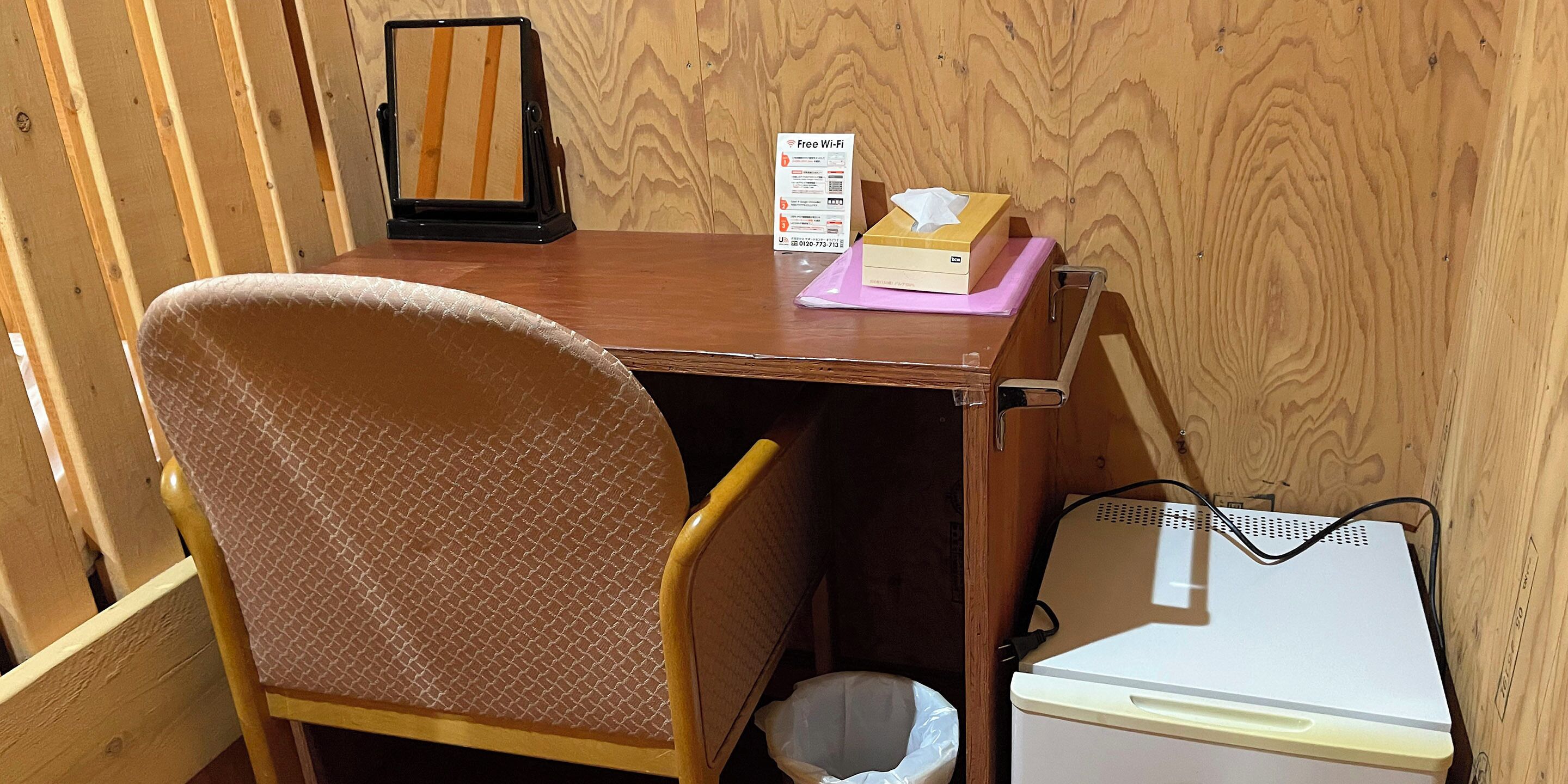 Un pequeño escritorio de madera con una silla, una caja de pañuelos, un cartel de Wi-Fi gratuito y un electrodoméstico blanco.