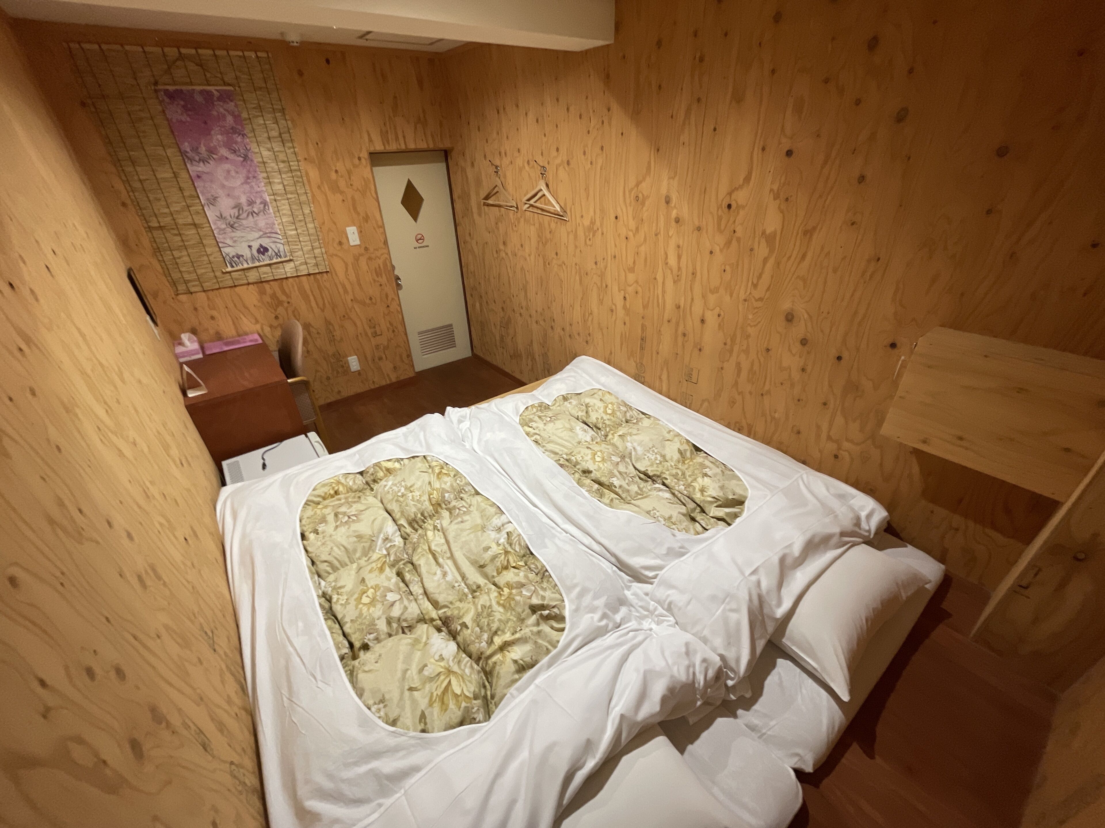 A small wooden room with two beds, a desk, and a wall-mounted shelf.
