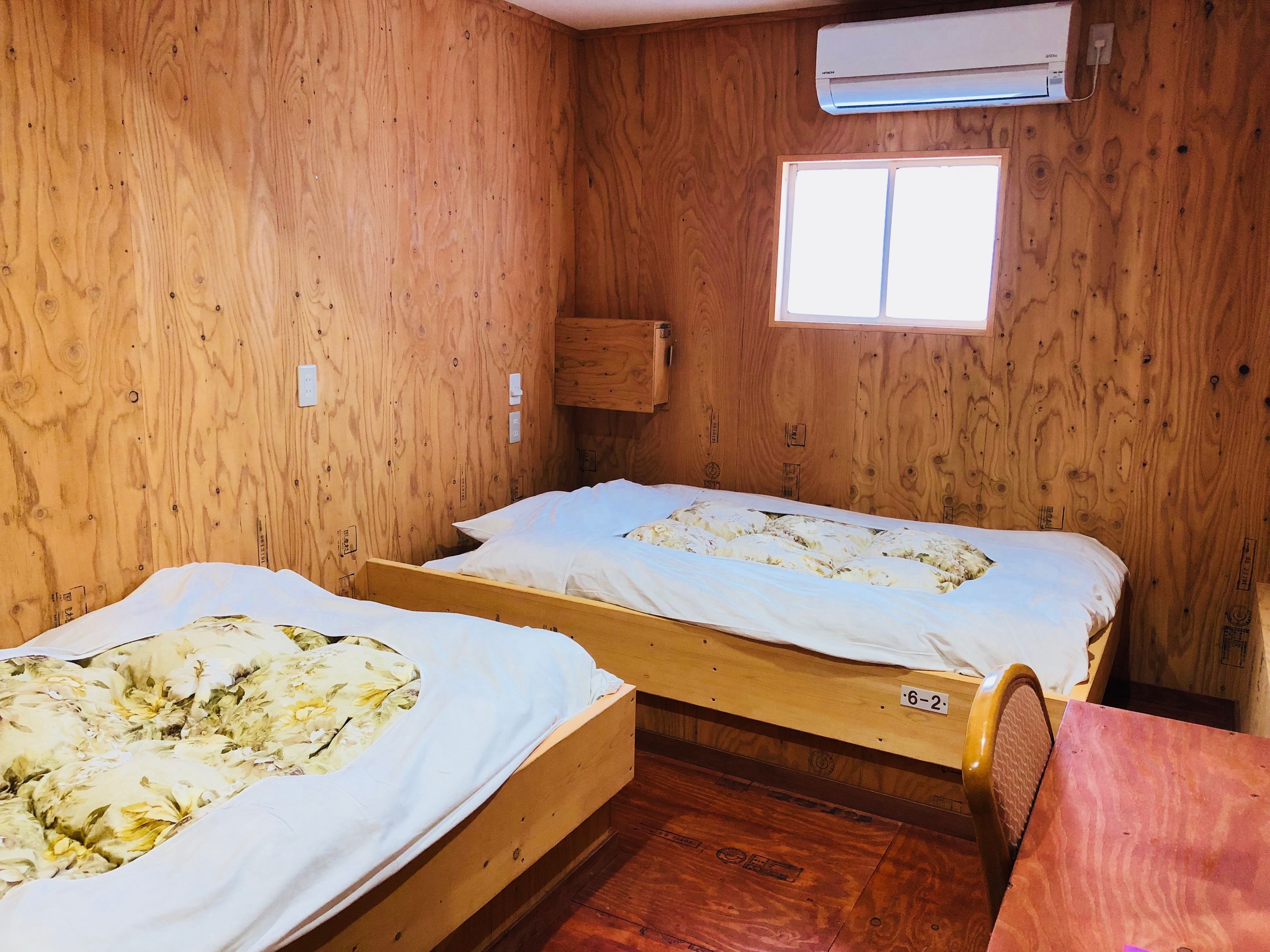 A small, wooden room with two single beds, a window, and an air conditioning unit.