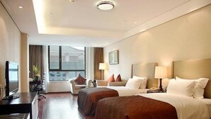 Ein Hotelzimmer mit zwei Betten, einem Schreibtisch mit Computer, einem Stuhl und einem Fenster mit Blick auf Gebäude.