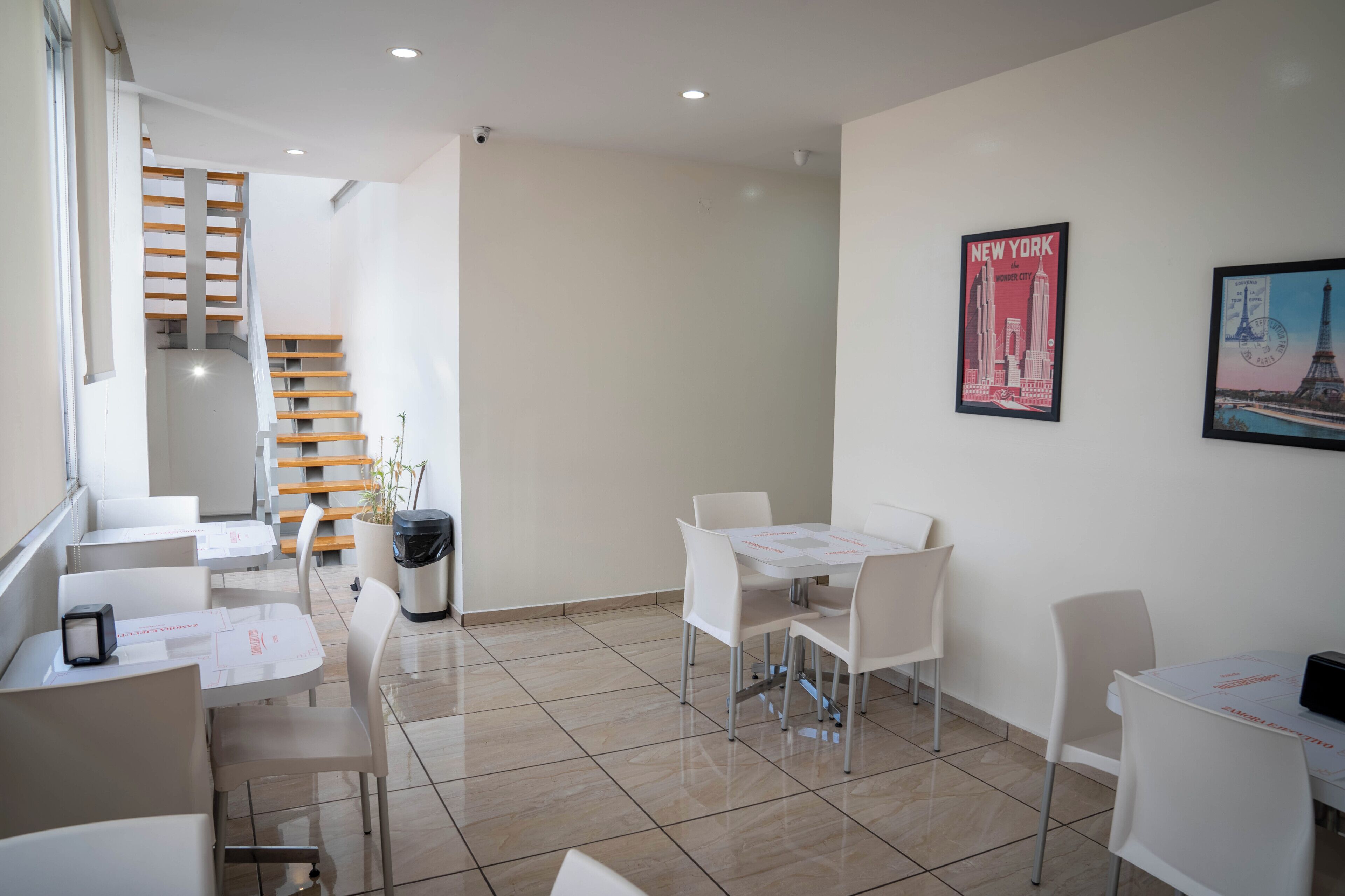 A hotel lobby with a staircase, white tables, and chairs, and two posters on the wall.