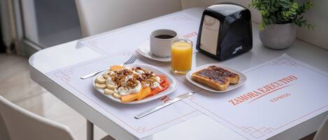 Una mesa de desayuno con un plato de comida, una taza de café, un vaso de jugo de naranja, un tostador y una planta en maceta.