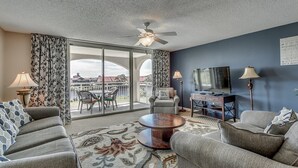 A living room with a sofa, armchairs, a coffee table, a TV, and a balcony with curtains. - Upscale Waterway View Condo, Onsite Golf Course | Yacht Club Villas - 3-303 (North Myrtle Beach)