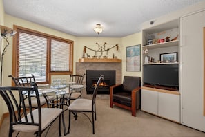 A living room with a fireplace, a dining table with chairs, a television on a stand, and a bookshelf.