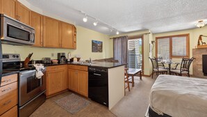 Cocina compacta con armarios de madera, microondas, estufa y lavavajillas. Cuenta con un comedor con mesa y sillas, además de una cama con cabecera.