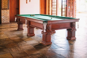 Una mesa de billar con superficie de fieltro verde, rodeada de suelo de madera y una pared de ladrillo.