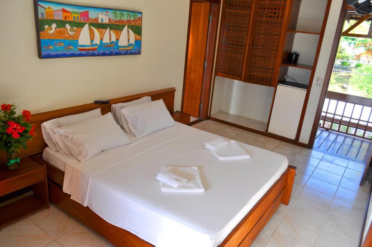 A neatly made bed with white linens, a wooden headboard, a nightstand with a vase of red flowers, and a painting of a coastal scene on the wall.