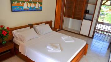 A neatly made bed with white linens, a wooden headboard, a nightstand with a vase of red flowers, and a painting of a coastal scene on the wall.