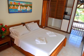 Una cama bien hecha con sábanas blancas, cabecera de madera, mesita de noche con un florero de flores rojas y un cuadro de una escena costera en la pared.