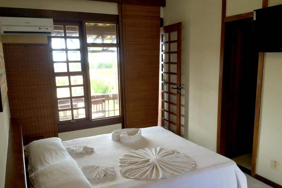 A neatly made bed with a white bedspread and decorative pillows, a window with a view of greenery, and a wooden door with a glass panel.