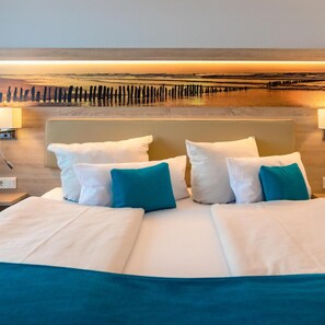 A hotel room with a bed, pillows, a headboard, and a wall-mounted artwork depicting a coastal scene.