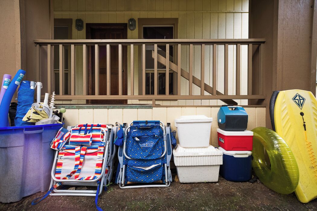 A collection of luggage and beach gear, including a blue suitcase, a striped beach chair, a white cooler, and a yellow surfboard, outside a building with a wooden staircase.