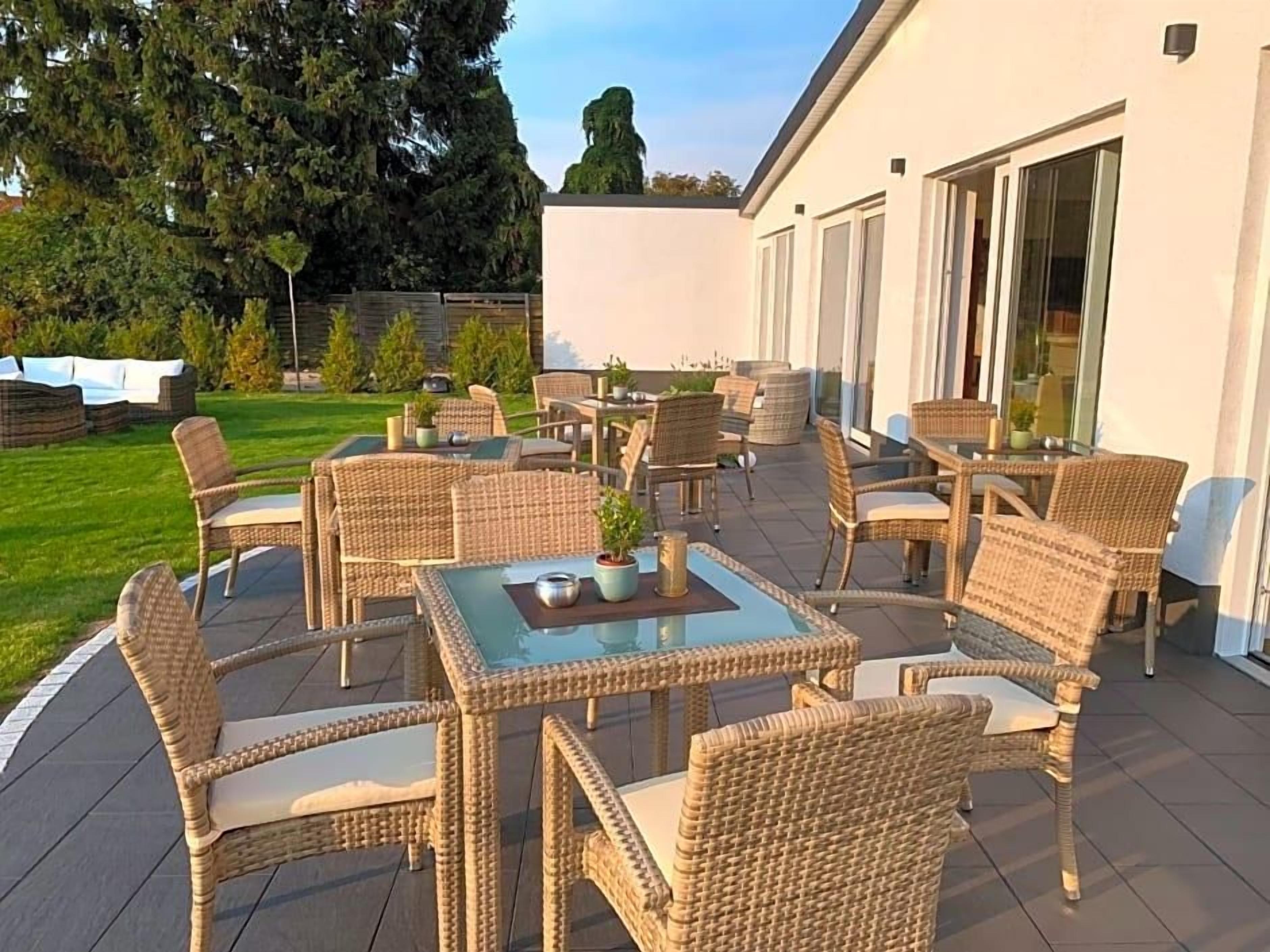 Un patio al aire libre con muebles de mimbre y mesas de vidrio, rodeado por un edificio blanco con grandes ventanales.