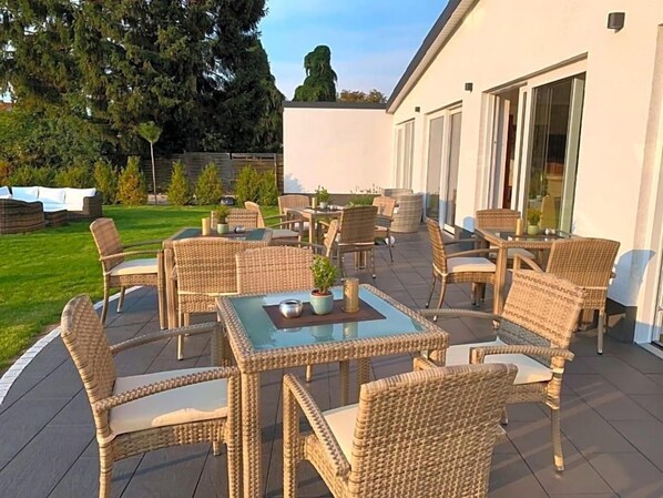An outdoor patio with wicker furniture and glass tables, surrounded by a white building with large windows.