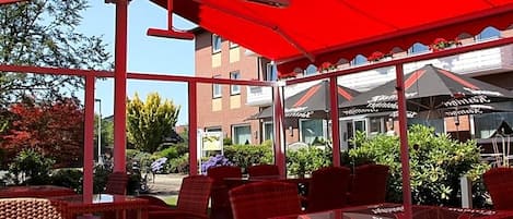 Ein Sitzbereich im Freien mit roten Stühlen und Tischen, einem roten Sonnensegel und Blick auf Gebäude im Hintergrund.