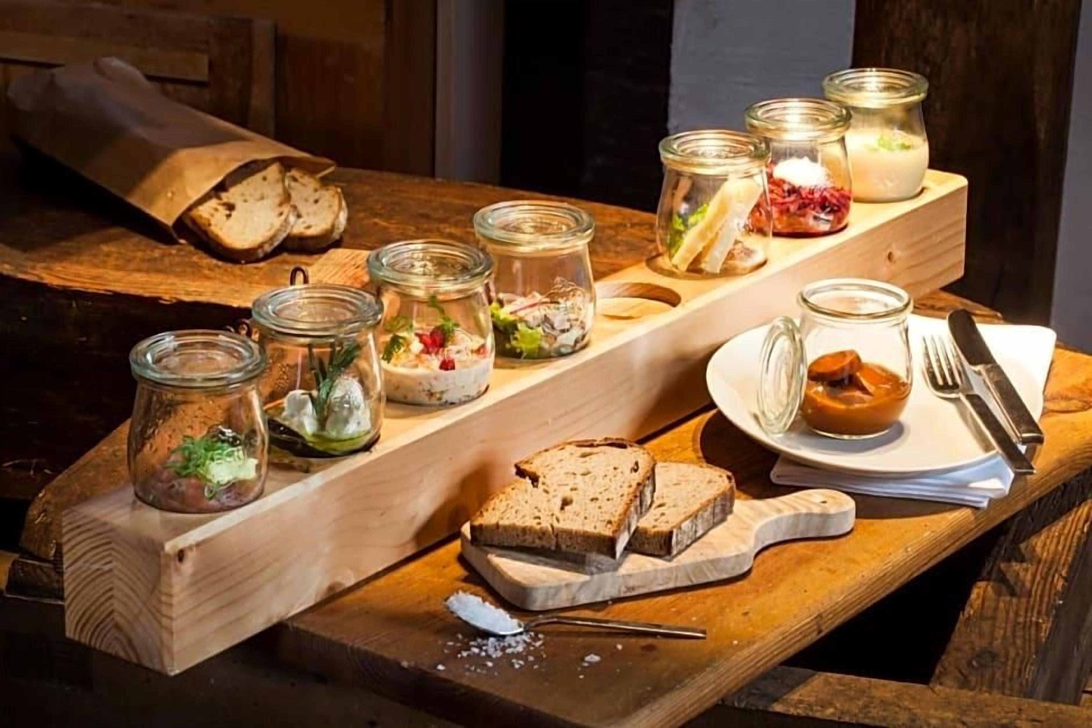 A wooden table with various food items and utensils, including bread, jars of pickles, and a plate with a fork and knife.