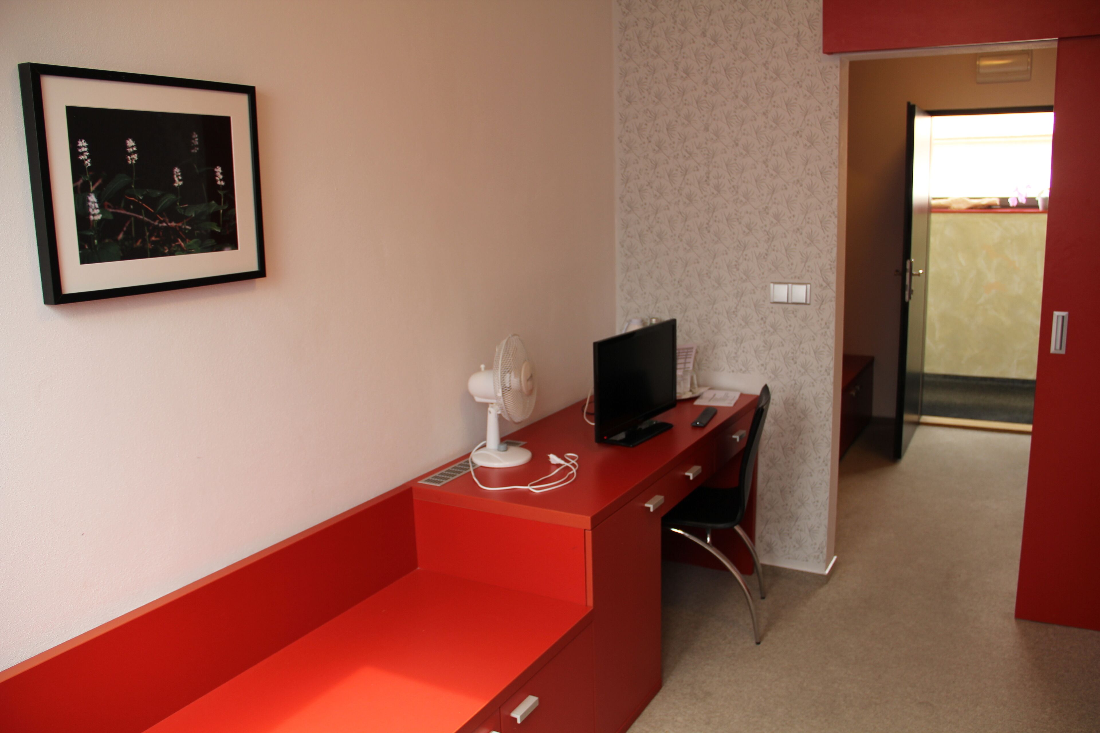 A room with a red desk, a framed picture on the wall, a fan, and a television.
