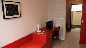 Une pièce avec un bureau rouge, un tableau encadré au mur, un ventilateur et un téléviseur.