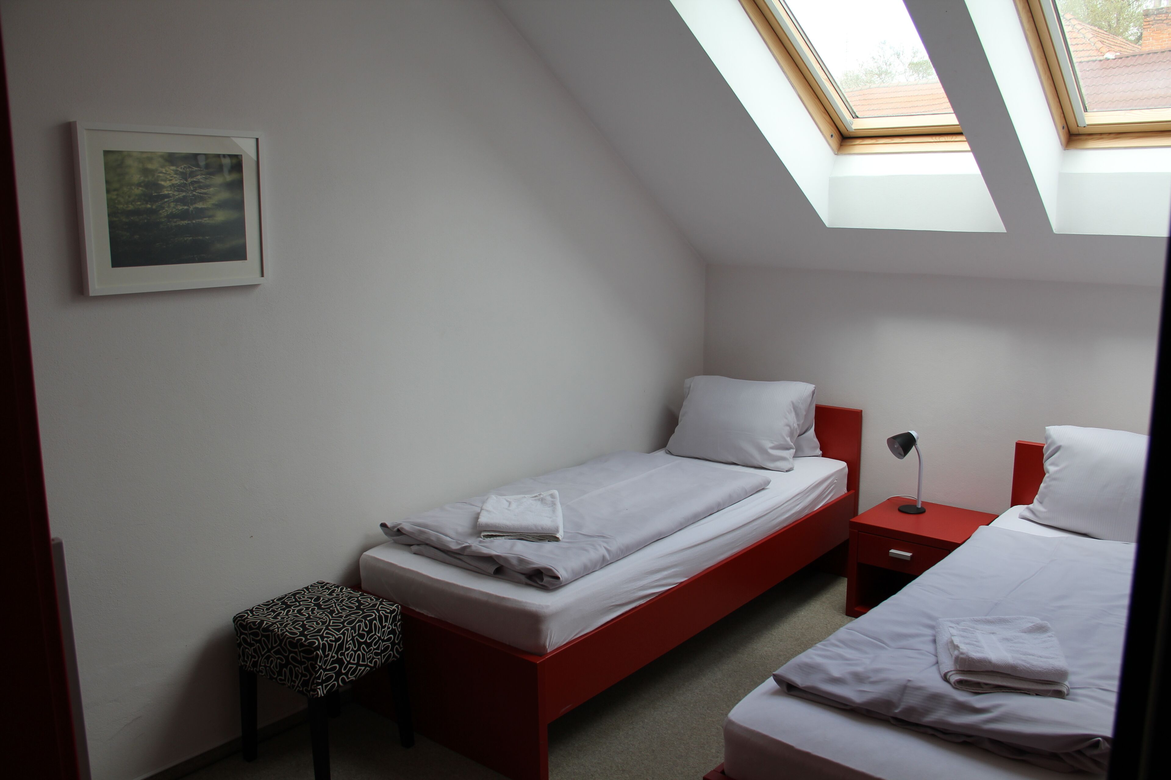 A bedroom with two beds, a skylight, a bedside table with a lamp, and a small stool.