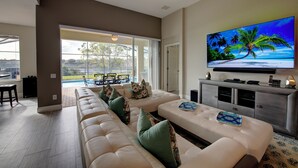 Una sala de estar moderna con un televisor de pantalla plana grande, un sofá modular blanco con cojines verdes y una puerta de vidrio que da a una zona de piscina.