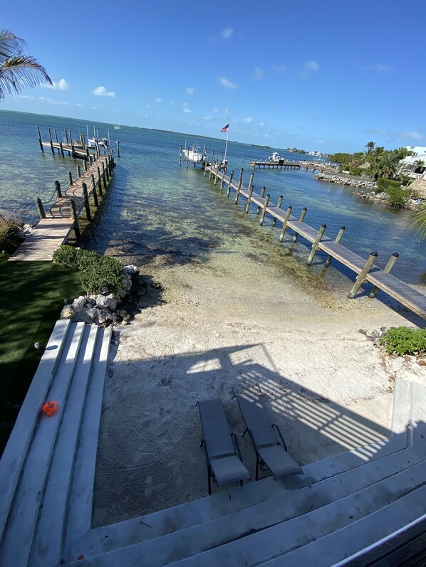 A wooden pier extending into clear blue water, with a sandy beach and a few lounge chairs.